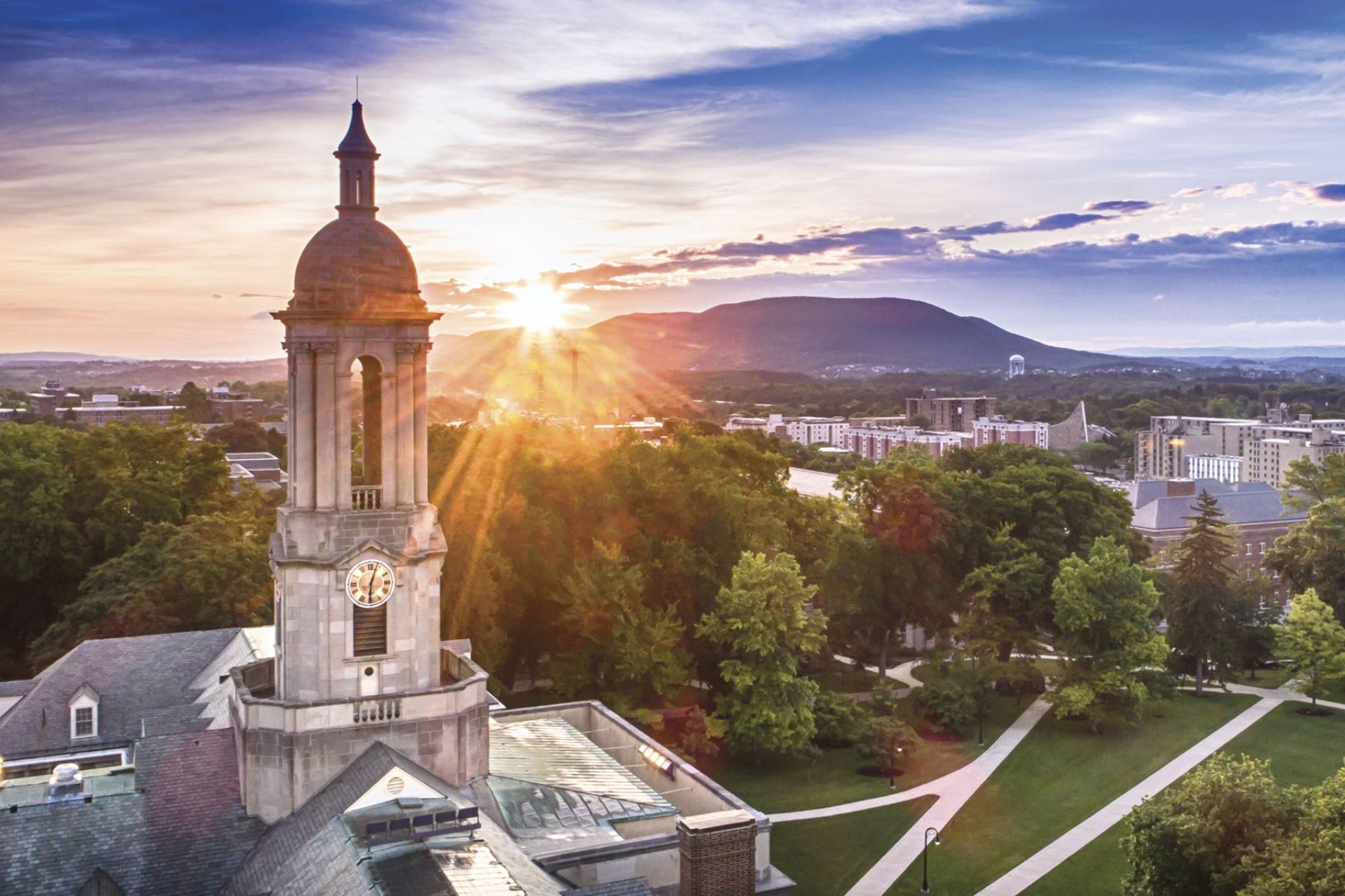 A clock tower building shot from the air by a drone sits in front of a wooded campus setting with the sun setting behind a hill in the background