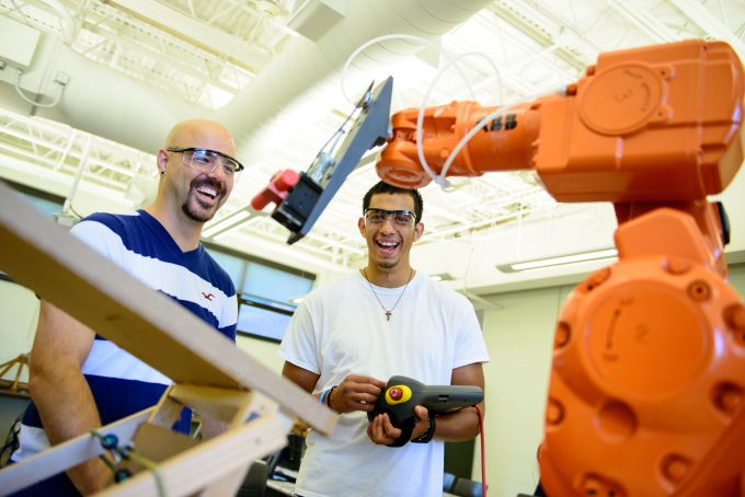 Two students wear safety glasses while working with an orange robotic arm in the foreground