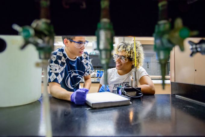 Two students are framed behind some chemistry equipment