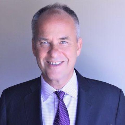 A man with neatly-trimmed graying hair smiles in a navy jacket and purple tie in front of a grayish-purple backdrop